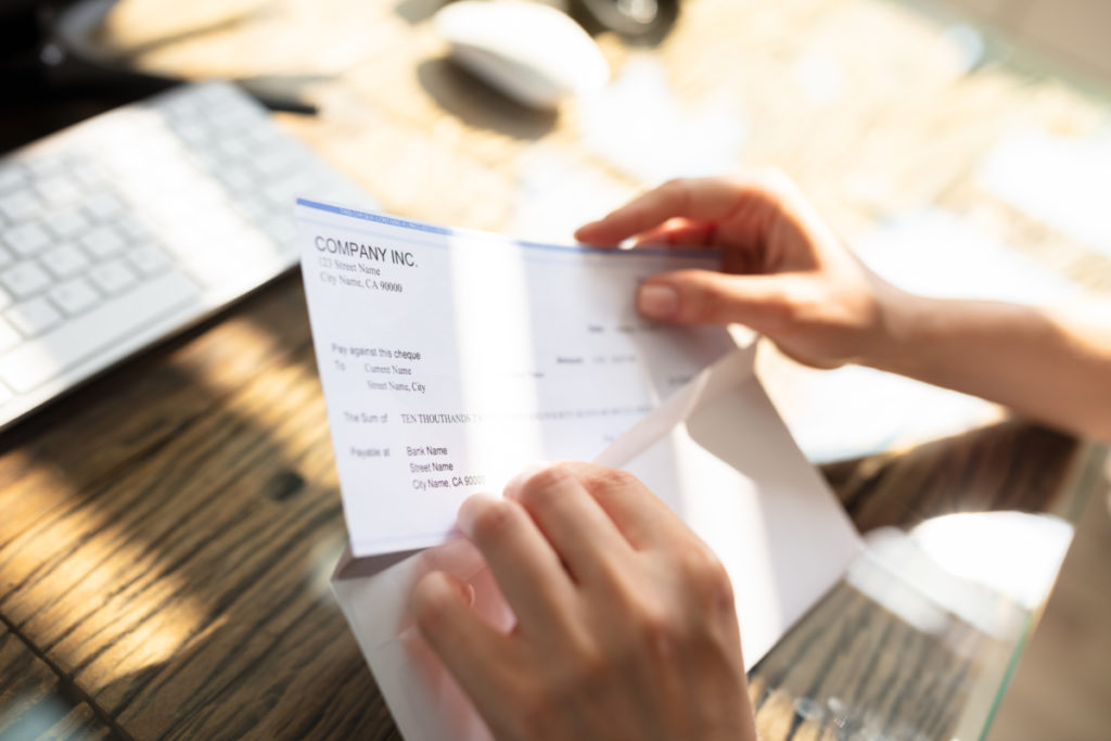 Close-up Of A Businessperson's Hand Opening Envelope With Paycheck Over Wooden Desk